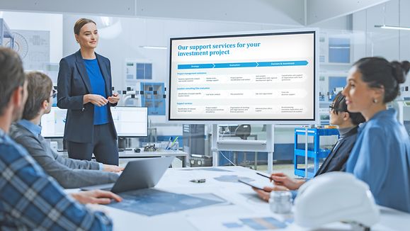 Modern Industrial Factory Meeting: Confident Female Engineer Uses Interactive Green Mock-up Screen Whiteboard, Makes Report to a Group of Engineers, Managers