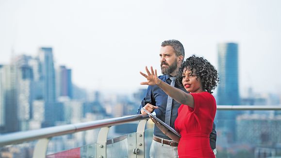 A portrait of businesspeople standing against London rooftop view, leaning on a railing and talking.